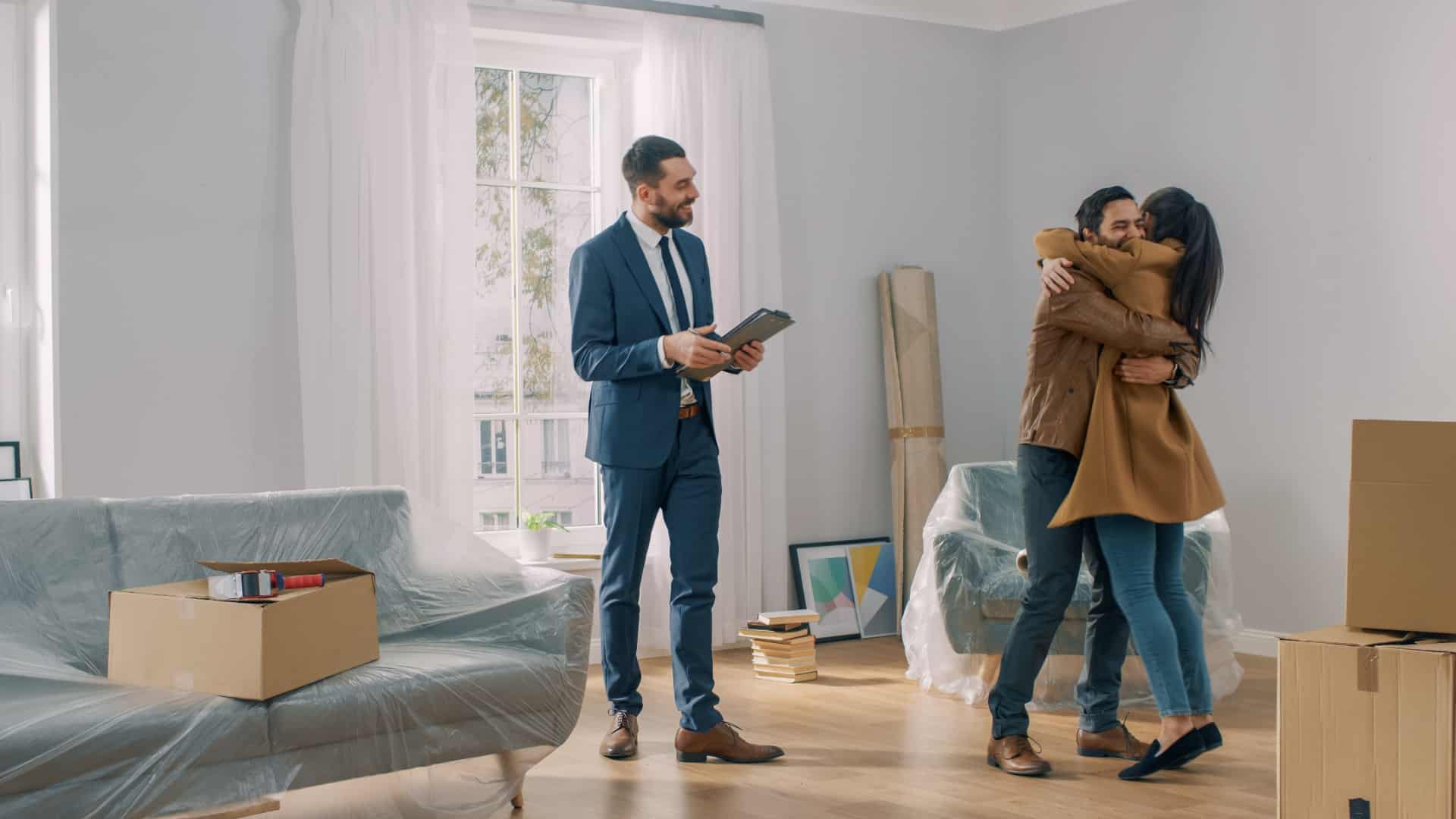 Real Estate Agent Shows Bright New Apartment to a Young Couple. Successful Young Couple Becoming Homeowners, Embraces and Hugs Each Other. Spacious Bright Home with Big Windows.