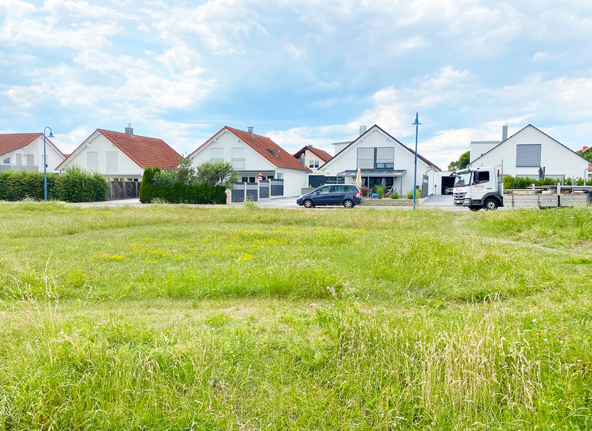 Eine ruhige Wohnstraße mit weißen Häusern, einem Auto und einem Lkw; blauer Himmel, grüne Wiese.